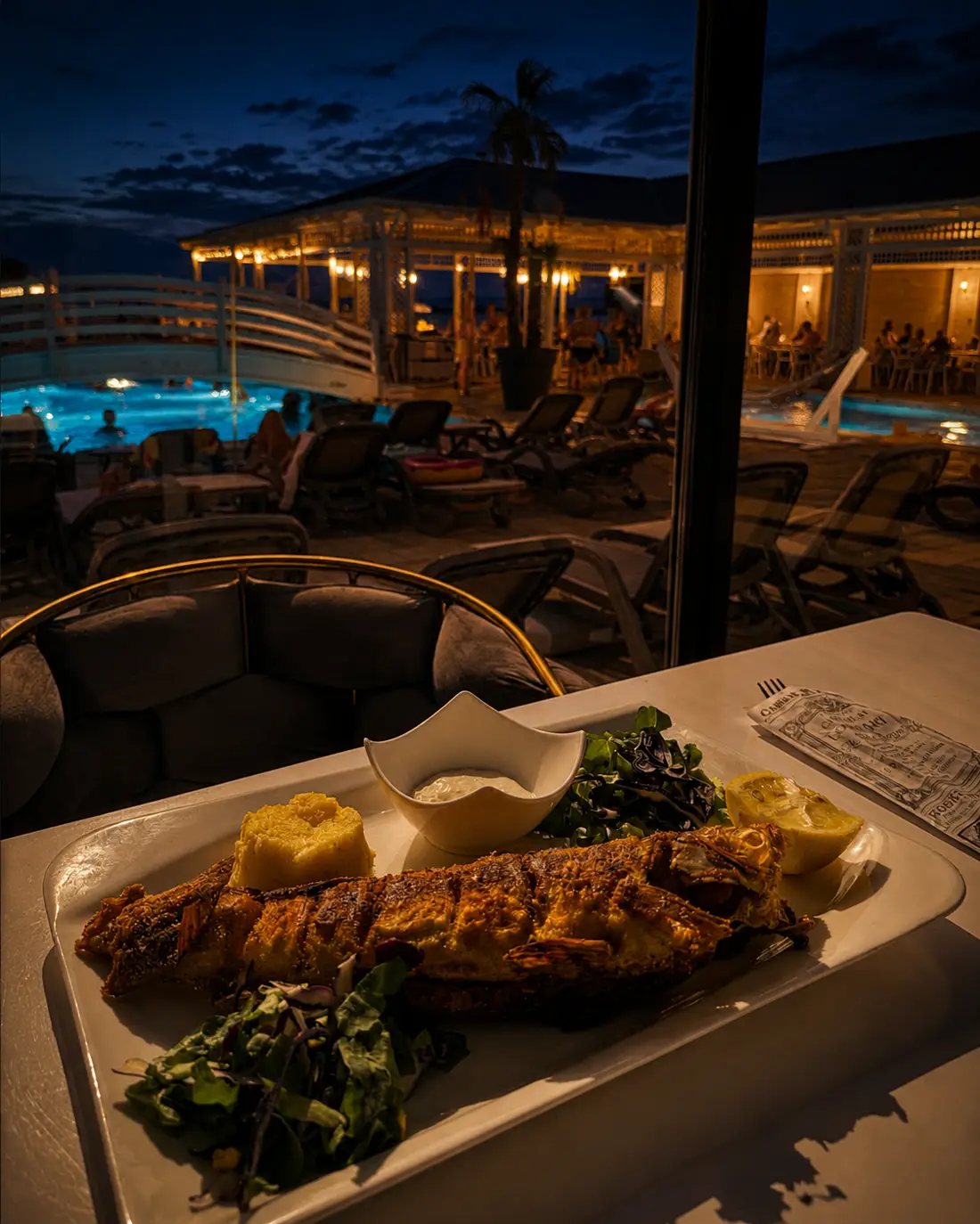Grilled fish with polenta and salad served at a window table overlooking the lit pool at night